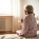 rear view senior sixty year old woman with gray hair holding mug washing down sleeping pill suffering from insomnia elderly retired female taking medicine with water sitting bedroom scaled 1