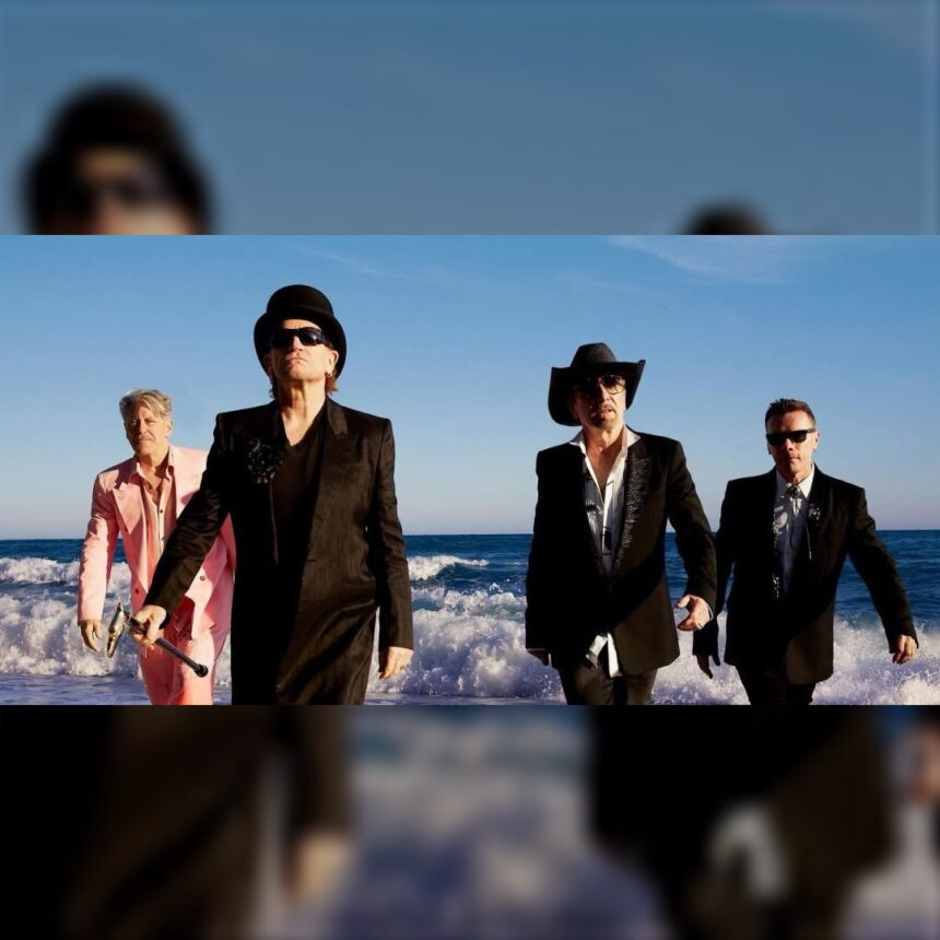 Os quatro integrantes do U2 caminham de terno na beira do mar, com ondas ao fundo