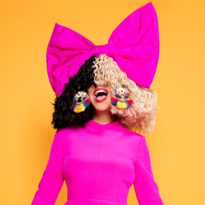 Sia com peruca metade preta e metade loira, laço rosa gigante na cabeça e vestido pink, em fundo laranja