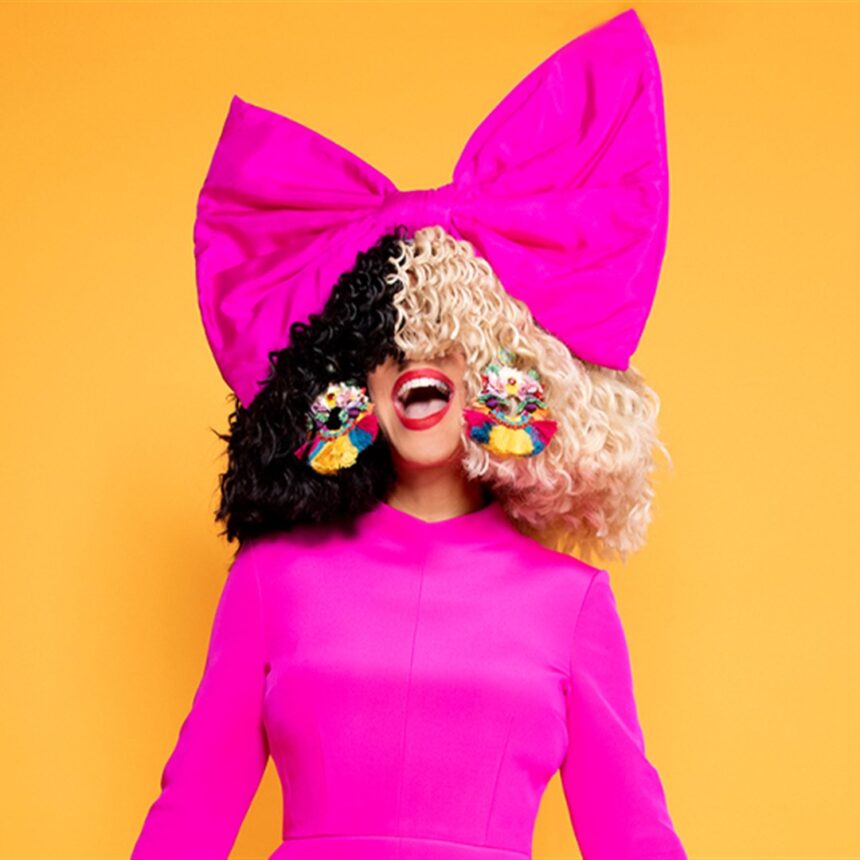 Sia com peruca metade preta e metade loira, laço rosa gigante na cabeça e vestido pink, em fundo laranja